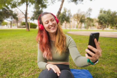 Parkta akıllı telefon kullanan kırmızı kulaklıklı, gülümseyen kadın..