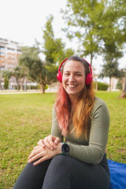 Parkta kırmızı kulaklıklı, mavi hasırda oturan memnun kadın..