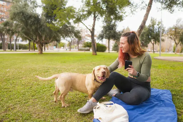 Parkta telefon tutarken köpeğini okşayan gülümseyen bir kadın..