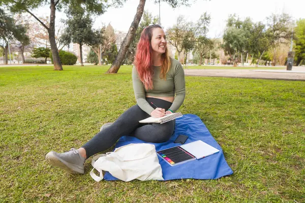 Parkta battaniyeye oturmuş gülümseyen bir kadın, bir defter çiziyor..