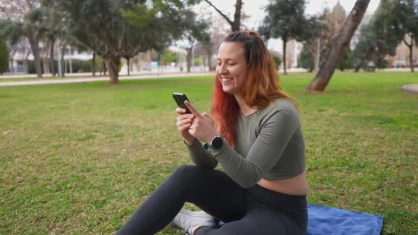 公園の青い毛布に座っている間 彼女のスマートフォンを使用して楽しい女性 — ストック動画
