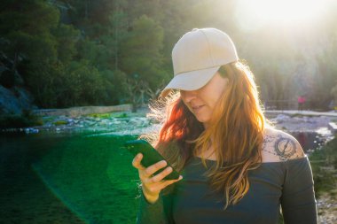Kadın telefonunu doğada kontrol ediyor, güneş ışığı içinden geçiyor..