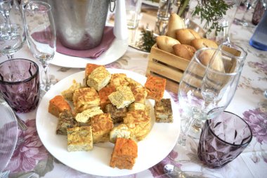 Beyaz tabakta çeşitli İspanyol omletleri, sepetinde ekmek ruloları ve sehpası var..