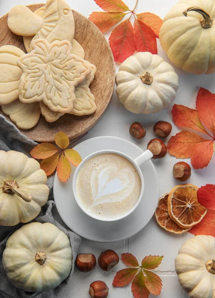 Cozy autumn composition. Hot coffee with cookies in a white cup  surrounded by autumn leaves and pumpkins on a white wooden background.