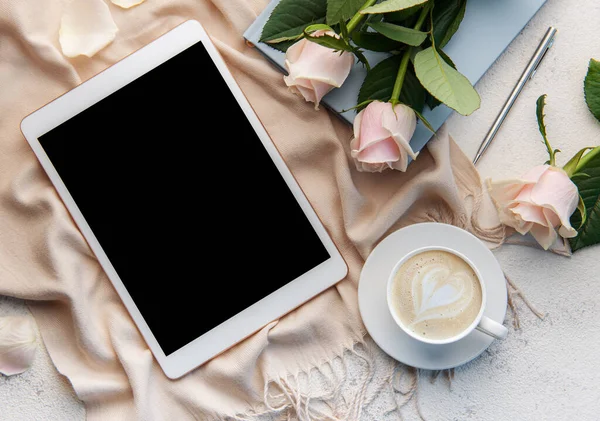 Minimalist workspace with digital tablet empty screen mockup, cozy scarf, a cup of coffee and roses flowers on concrete background. Top view, flat lay