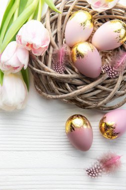 Nest with Easter eggs painted pink and golden colors and pink tulips on a white wooden background. Top view. Space for text.