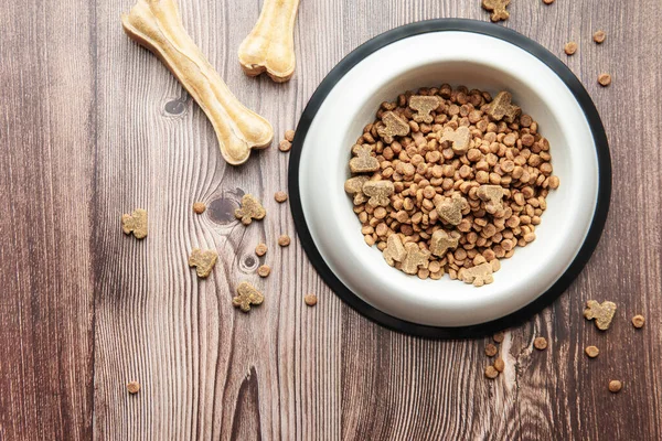 A bowl of dog food and treats in the form of bones on a wooden floor. Dry food in granules.