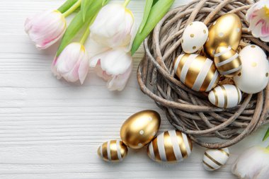 Nest with Easter eggs painted golden colors and pink tulips on a white wooden background. Top view. Space for text.