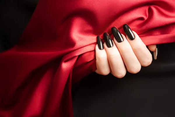 Hands of young girl with  black manicure on nails holding red satin cloth on black background