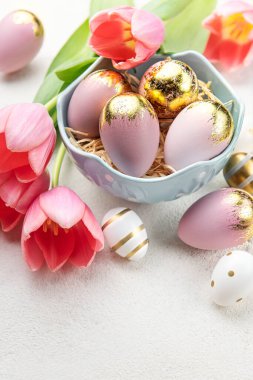 Bowl with Easter eggs painted gold and pastel colors and pink tulips on a concrete background. Top view. Space for text.
