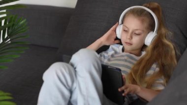 Kulaklıklı ve dijital tabletli müzik dinleyen küçük bir kız. Tablet oyunu, kulaklıklar ve rahatlama çocuk oyunları, eğlenmek veya online oyunlar oynamak veya müzik dinlemek
