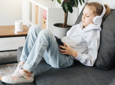 Kanepede elektronik tabletle uzanmış küçük bir kız beyaz kulaklıkla müzik dinliyor. Eğitim için kablosuz aygıtlar kullanılıyor.
