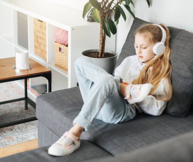 Kanepede elektronik tabletle uzanmış küçük bir kız beyaz kulaklıkla müzik dinliyor. Eğitim için kablosuz aygıtlar kullanılıyor.