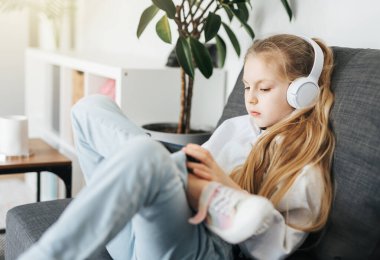 Kanepede elektronik tabletle uzanmış küçük bir kız beyaz kulaklıkla müzik dinliyor. Eğitim için kablosuz aygıtlar kullanılıyor.