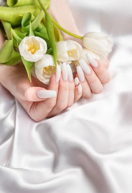 Yay tırnağı dizaynlı bir kadın eli. Beyaz ojeli manikür. Mükemmel manikürlü kadın model el beyaz ipek arka planda beyaz lale çiçekleri tutun