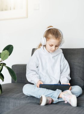 Kanepede elektronik tabletle uzanmış küçük bir kız beyaz kulaklıkla müzik dinliyor. Eğitim için kablosuz aygıtlar kullanılıyor.