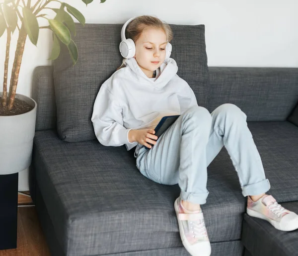 小さな女の子は電子タブレットとソファの上に横たわって白いヘッドフォンで音楽を聴く 教育用無線装置の使用 — ストック写真