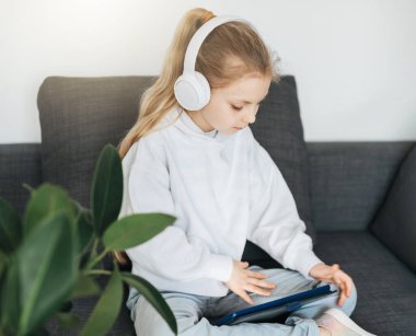 Kanepede elektronik tabletle uzanmış küçük bir kız beyaz kulaklıkla müzik dinliyor. Eğitim için kablosuz aygıtlar kullanılıyor.