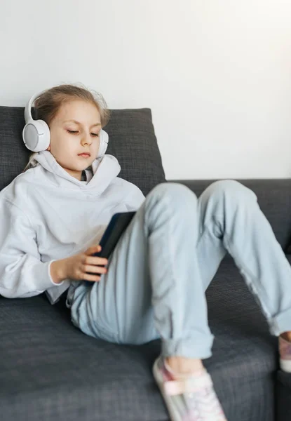 Pieni Tyttö Makaa Sohvalla Elektroninen Tabletti Kuuntelee Musiikkia Valkoiset Kuulokkeet — kuvapankkivalokuva
