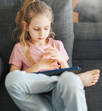 Elektronik tabletle kanepede yatan küçük bir kız. Eğitim için kablosuz aygıtlar kullanılıyor.