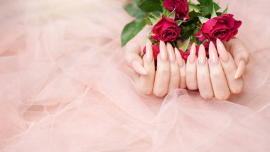 Zarif pastel pembe doğal manikür. Pembe ipek arka planda gülleri olan kadın elleri.