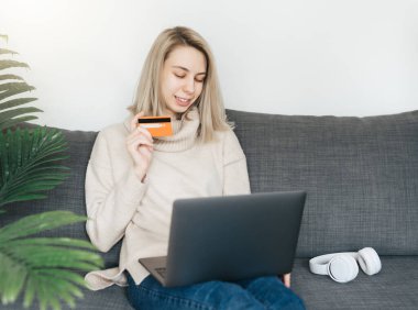 Kredi kartı tutan ve dizüstü bilgisayar kullanan genç bir kadın. İnternette çalışan bir kız. Çevrimiçi alışveriş, internet bankacılığı, evden çalışma 