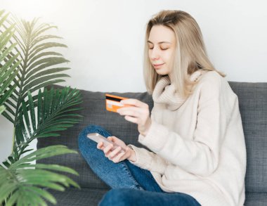 Akıllı telefon ve bankacılık kredi kartı tutan genç bir kadın, evde internet üzerinden alışveriş yapan mutlu bir kadın, internet mağazasından mal veya hizmet satın alan mutlu bir kadın..