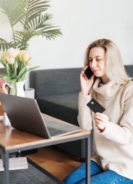 Kredi kartı tutan ve dizüstü bilgisayar kullanan genç bir kadın. Evde çalışan bir kadın. Çevrimiçi alışveriş, internet bankacılığı, ev konseptinden çalışmak
