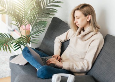 Evde dizüstü bilgisayar kullanan genç bir kadın oturma odasında rahat bir şekilde oturuyor.. 