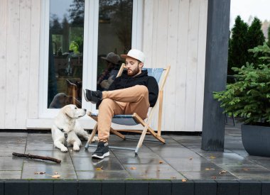 Genç bir adam bir golden retriever ile kır evinin yakınında dinleniyor.. 