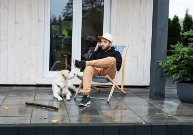 Genç bir adam bir golden retriever ile kır evinin yakınında dinleniyor.. 