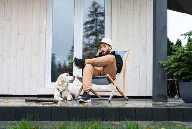 Genç bir adam bir golden retriever ile kır evinin yakınında dinleniyor.. 