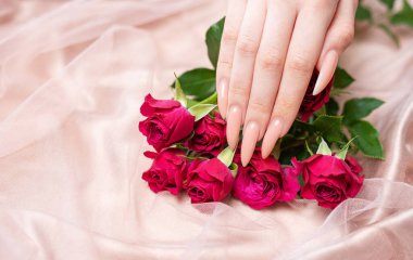 Zarif pastel pembe doğal manikür. Pembe ipek arka planda gülleri olan kadın elleri.