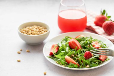 Canlı bir tabak çilek roka salatası, kavrulmuş çam fıstığıyla süslenmiş, beyaz bir tabakta sunuluyor, açık gri bir arka planda. Yeşilliklerin tazeliği ve çileklerin olgunluğu sağlıklı ve besleyici bir yemek seçeneği sunuyor..