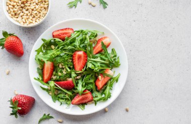 Canlı bir tabak çilek roka salatası, kavrulmuş çam fıstığıyla süslenmiş, beyaz bir tabakta sunuluyor, açık gri bir arka planda. Yeşilliklerin tazeliği ve çileklerin olgunluğu sağlıklı ve besleyici bir yemek seçeneği sunuyor..