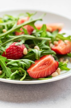 Canlı bir tabak çilek roka salatası, kavrulmuş çam fıstığıyla süslenmiş, beyaz bir tabakta sunuluyor, açık gri bir arka planda. Yeşilliklerin tazeliği ve çileklerin olgunluğu sağlıklı ve besleyici bir yemek seçeneği sunuyor..