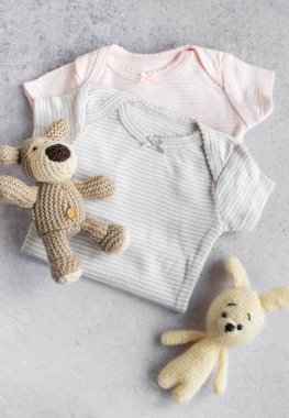 Gri arka planda bebek kıyafetleri. Yavru örülmüş köpek ve tavşan oyuncağı çocuklar için tulumların yanında. 