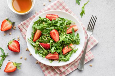 Canlı bir tabak çilek roka salatası, kavrulmuş çam fıstığıyla süslenmiş, beyaz bir tabakta sunuluyor, açık gri bir arka planda. Yeşilliklerin tazeliği ve çileklerin olgunluğu sağlıklı ve besleyici bir yemek seçeneği sunuyor..