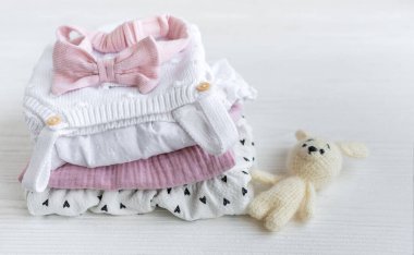 Bebek kıyafetleri yığını, pembe saç bandı ve beyaz tahta arka planda örülmüş oyuncak tavşan.