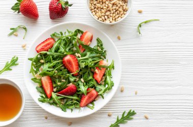 Canlı bir tabak çilek roka salatası, kavrulmuş çam fıstığıyla süslenmiş, beyaz bir tabakta sunuluyor, açık gri bir arka planda. Yeşilliklerin tazeliği ve çileklerin olgunluğu sağlıklı ve besleyici bir yemek seçeneği sunuyor..