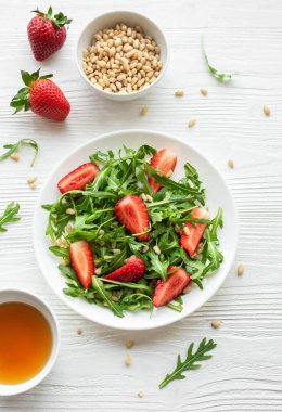 Canlı bir tabak çilek roka salatası, kavrulmuş çam fıstığıyla süslenmiş, beyaz bir tabakta sunuluyor, açık gri bir arka planda. Yeşilliklerin tazeliği ve çileklerin olgunluğu sağlıklı ve besleyici bir yemek seçeneği sunuyor..