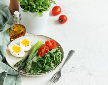 Sağlıklı bir bahar salatası beyaz bir masada, içinde taze roka yapraklarının yanında dilimlenmiş yumurta, canlı yeşil kuşkonmaz ve olgun vişne domatesleri bulunur.. 