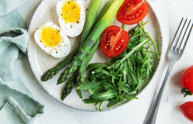 Sağlıklı bir bahar salatası beyaz bir masada, içinde taze roka yapraklarının yanında dilimlenmiş yumurta, canlı yeşil kuşkonmaz ve olgun vişne domatesleri bulunur..