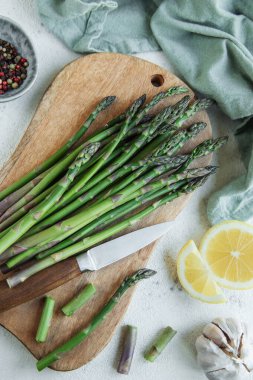 Fresh green asparagus lies on a wooden cutting board alongside a kitchen knife, with slices of lemon and garlic cloves nearby. The cutting board is on a tabletop adorned with a green cloth, and peppercorns are visible in the background, suggesting a 