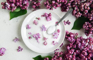 Şekerle dolu beyaz bir kase hem kasenin içinde hem de açık renkli bir yüzeye saçılmış leylak çiçekleriyle süslenir. Klozetin içinde bir kaşık var. Bu ayar yemek pişirmek ya da servis etmek için kullanılabilir..