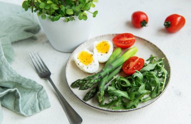 Sağlıklı bir bahar salatası beyaz bir masada, içinde taze roka yapraklarının yanında dilimlenmiş yumurta, canlı yeşil kuşkonmaz ve olgun vişne domatesleri bulunur..
