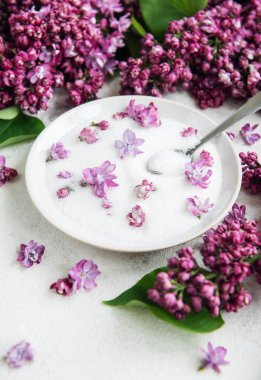 Şekerle dolu beyaz bir kase hem kasenin içinde hem de açık renkli bir yüzeye saçılmış leylak çiçekleriyle süslenir. Klozetin içinde bir kaşık var. Bu ayar yemek pişirmek ya da servis etmek için kullanılabilir..