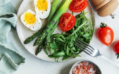 Sağlıklı bir bahar salatası beyaz bir masada, içinde taze roka yapraklarının yanında dilimlenmiş yumurta, canlı yeşil kuşkonmaz ve olgun vişne domatesleri bulunur..