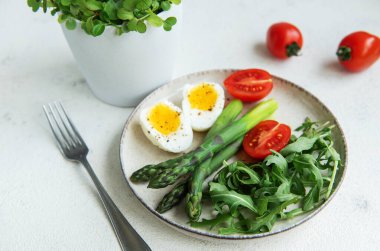 Sağlıklı bir bahar salatası beyaz bir masada, içinde taze roka yapraklarının yanında dilimlenmiş yumurta, canlı yeşil kuşkonmaz ve olgun vişne domatesleri bulunur..