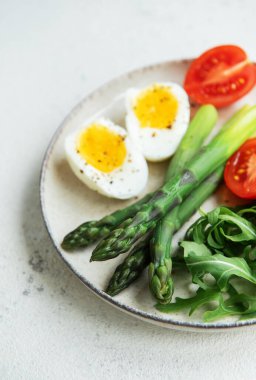 Sağlıklı bir bahar salatası beyaz bir masada, içinde taze roka yapraklarının yanında dilimlenmiş yumurta, canlı yeşil kuşkonmaz ve olgun vişne domatesleri bulunur..